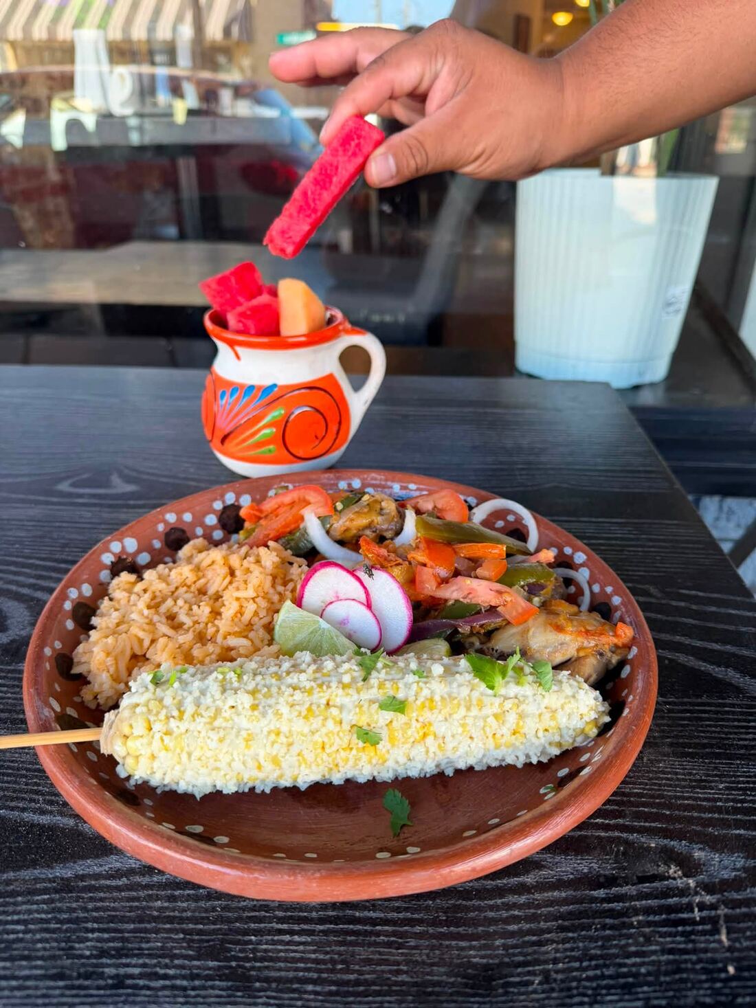 Elote and grilled shrimp on a clay plate, with salsa being poured from a painted jug